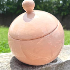 May include: A spherical ceramic jar with a smooth, glossy, light peach-colored finish. The jar features a matching rounded lid with a small, knob-like handle at the top, creating a uniform, minimalist, and clean aesthetic for this container.