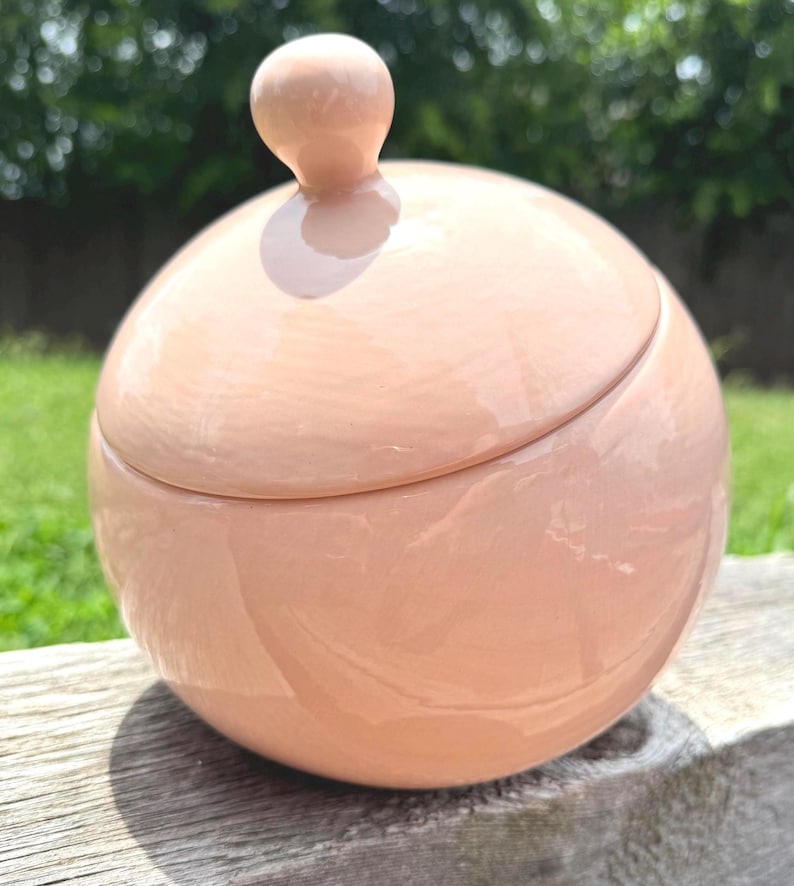 May include: A spherical ceramic jar with a smooth, glossy, light peach-colored finish. The jar features a matching rounded lid with a small, knob-like handle at the top, creating a uniform, minimalist, and clean aesthetic for this container.