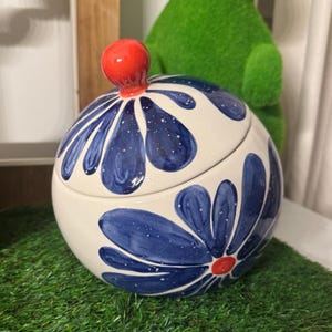 May include: A round ceramic jar with a lid, featuring a cream-colored base decorated with large, hand-painted blue flower petals that have white speckles. The lid is topped with a small, bright orange spherical knob, and the center of the flower on the jar body also features a small orange dot.