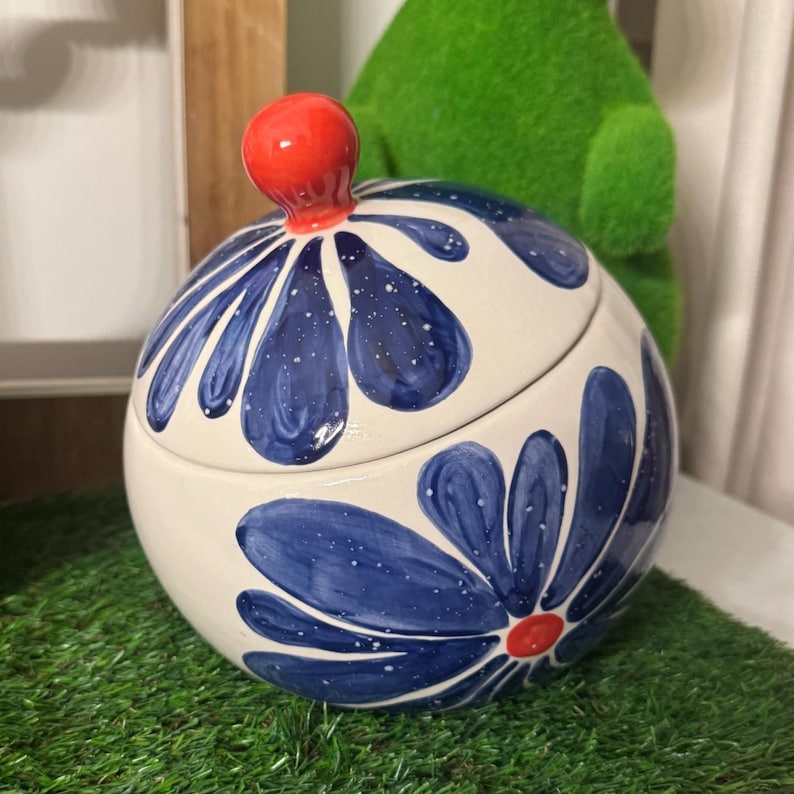 May include: A round ceramic jar with a lid, featuring a cream-colored base decorated with large, hand-painted blue flower petals that have white speckles. The lid is topped with a small, bright orange spherical knob, and the center of the flower on the jar body also features a small orange dot.