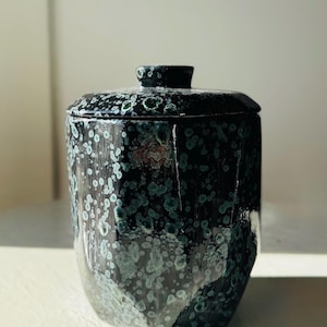 May include: A cylindrical ceramic jar with a matching lid, featuring a glossy black glaze decorated with a speckled pattern of light blue and teal spots. The vessel has a faceted, multi-sided exterior surface and a small knob handle on top.