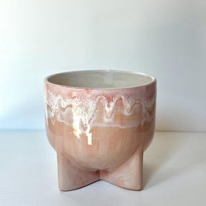 May include: A ceramic planter with a rounded body and three short, sturdy feet. The exterior features a smooth, light peach-coloured glaze on the lower portion, transitioning to a textured, drippy white and soft pink glaze around the upper rim.