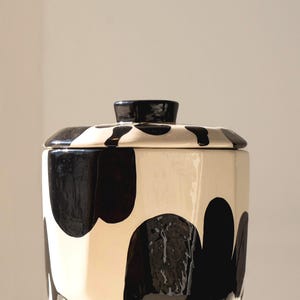 Puede incluir: Un tarro de cerámica con tapa, con base color crema y grandes formas negras irregulares. La tapa tiene un pequeño asa redondeada. El diseño es moderno y minimalista, adecuado para la cocina.