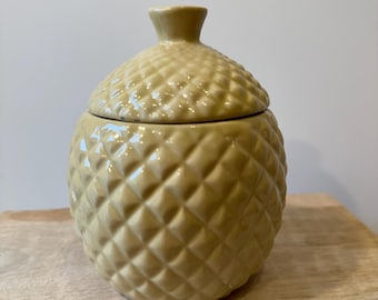 May include: A pale yellow ceramic jar with a lid, featuring a textured, diamond-shaped pattern across its entire surface. The container has a rounded, bulbous shape and a small, knob-like handle on top of the lid, with a glossy finish.