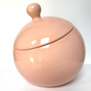 May include: A spherical ceramic jar with a smooth, glossy light pink finish. The container features a matching rounded lid topped with a small, ball-shaped handle. The surface reflects light, highlighting the curved, uniform shape of the vessel.