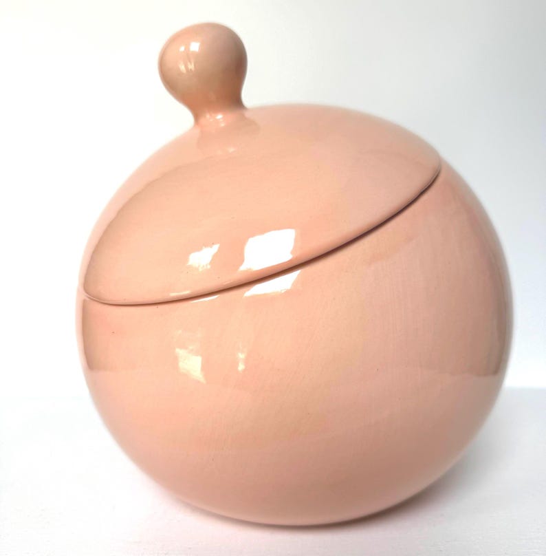 May include: A spherical ceramic jar with a smooth, glossy light pink finish. The container features a matching rounded lid topped with a small, ball-shaped handle. The surface reflects light, highlighting the curved, uniform shape of the vessel.