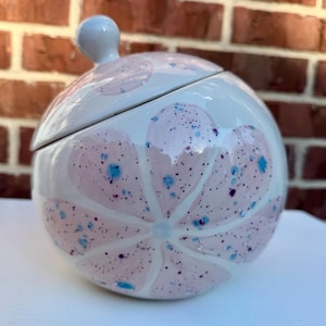 May include: A round ceramic lidded jar with a glossy white finish, featuring a large painted pink flower design. The surface is decorated with small, scattered blue and dark purple speckles throughout the petals and the white background.