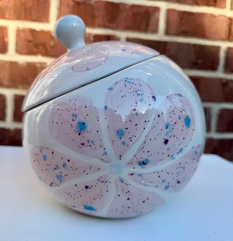 May include: A round ceramic lidded jar with a glossy white finish, featuring a large painted pink flower design. The surface is decorated with small, scattered blue and dark purple speckles throughout the petals and the white background.