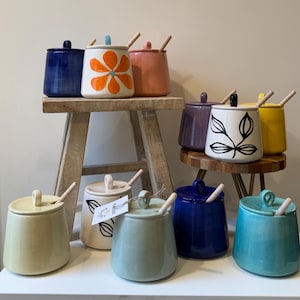 Könnte beinhalten: Eine Sammlung farbenfroher Keramikgläser mit Deckeln und Holzlöffeln. Die Gläser sind in verschiedenen Farben erhältlich, darunter Blau, Weiß mit einer orangefarbenen Blume, Rosa, Lila, Weiß mit einem Blattmuster, Gelb, Hellgrün, Creme und Türkis.