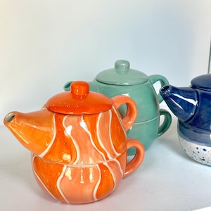 May include: A stackable ceramic teapot and cup set featuring a bright orange marbled pattern with white swirls and a speckled lid. Behind it sits a solid sage green teapot and cup set, and to the right is a dark blue and white speckled teapot.