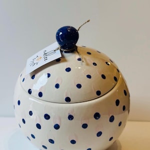 May include: A round ceramic jar with a lid, featuring a cream-colored base decorated with dark blue polka dots and faint pink brushstrokes. A dark blue spherical knob sits on top of the lid, which has a white paper tag labeled Nikkee Pottery.