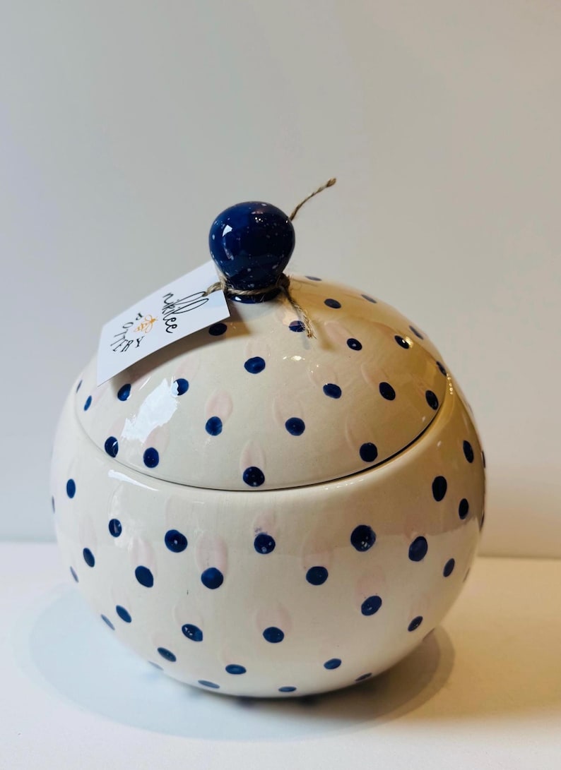 May include: A round ceramic jar with a lid, featuring a cream-colored base decorated with dark blue polka dots and faint pink brushstrokes. A dark blue spherical knob sits on top of the lid, which has a white paper tag labeled Nikkee Pottery.