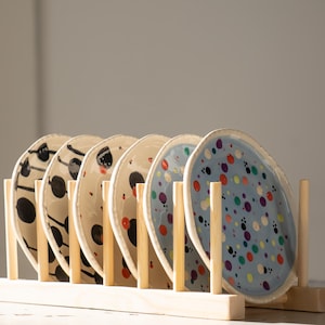 Może przedstawiać: Zestaw sześciu ceramicznych talerzy z unikalnymi, ręcznie malowanymi wzorami, prezentowanych w drewnianym stojaku na talerze. Talerze mają czarne, czerwone i niebieskie wzory na kremowym i jasnoniebieskim tle. Stojak jest wykonany z jasnego drewna.