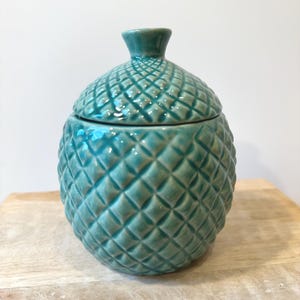 May include: A teal ceramic jar with a lid, resembling a pineapple. The jar has a textured, diamond-patterned surface and a small knob on the lid. The jar is placed on a wooden surface.