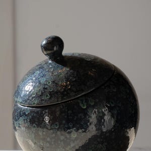 May include: A spherical ceramic jar with a lid featuring a small, rounded knob handle. The surface has a glossy, dark finish with mottled speckles in shades of deep blue, black, and hints of teal, creating a textured, multi-tonal appearance.