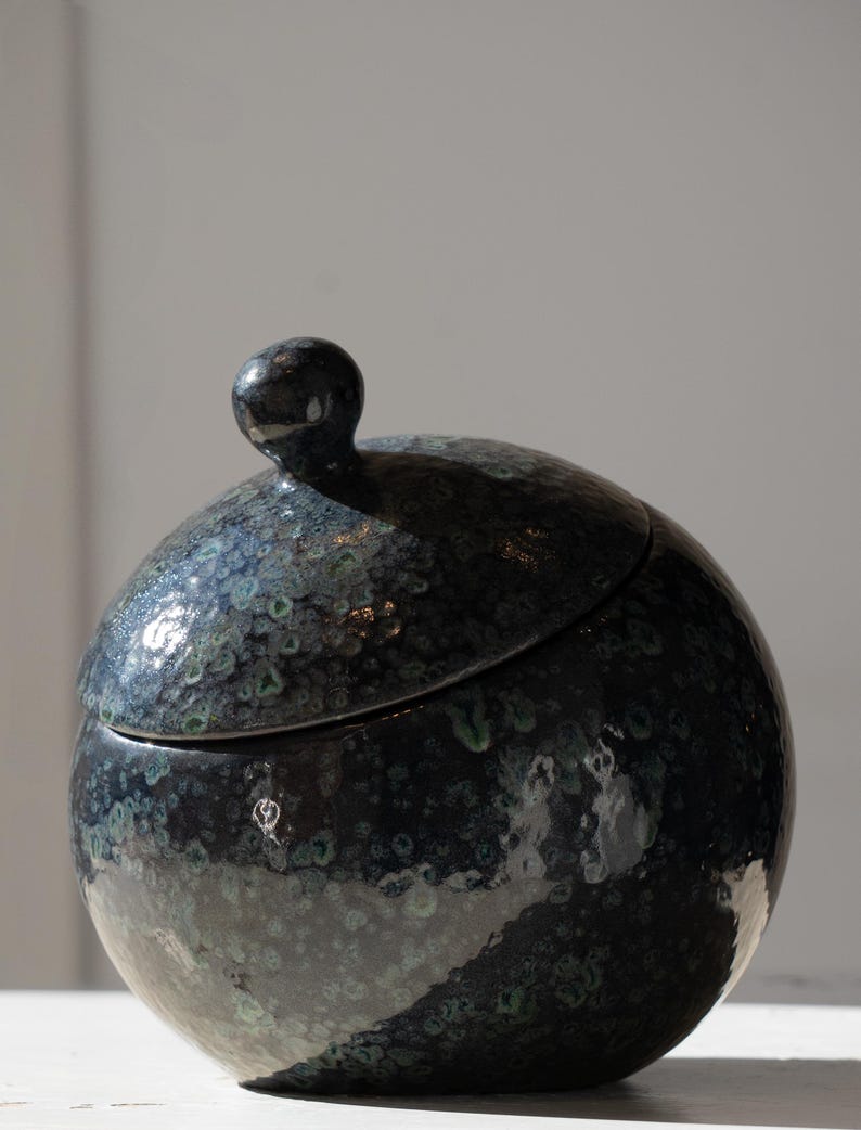 May include: A spherical ceramic jar with a lid featuring a small, rounded knob handle. The surface has a glossy, dark finish with mottled speckles in shades of deep blue, black, and hints of teal, creating a textured, multi-tonal appearance.
