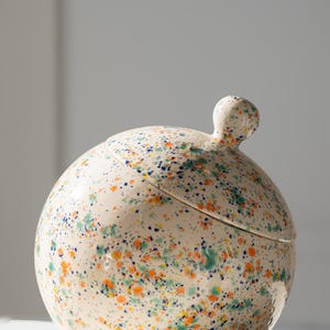 May include: A spherical ceramic jar with a matching lid featuring a small round knob handle. The surface is off-white and covered in a speckled pattern of small orange, teal, and dark blue paint splatters across the entire exterior.