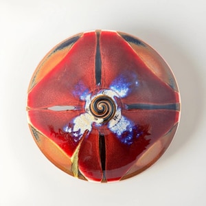 May include: A circular ceramic bowl featuring a glossy glaze in shades of deep red, burnt orange, and black. The center displays a prominent dark spiral pattern surrounded by mottled blue and white accents, with four distinct radiating sections.