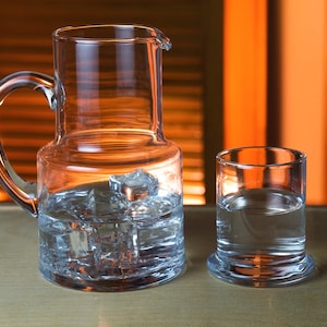 May include: A clear glass pitcher with a handle and a matching glass, both filled with water and ice cubes. The pitcher is on a wooden surface, with an orange background.
