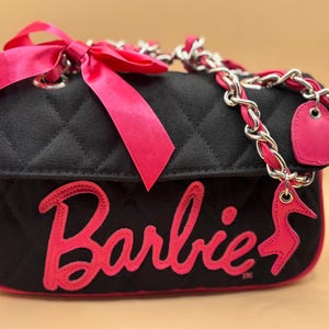 May include: A black quilted handbag featuring the pink embroidered text Barbie and a matching pink high heel shoe charm. The bag includes a silver chain strap interwoven with pink ribbon, a large pink bow on the side, and a pink heart-shaped tag.