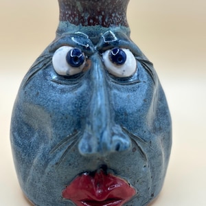 May include: A ceramic face jug with a blue glazed finish, featuring a sculpted human-like face with prominent red lips, a long nose, and two white eyes with dark blue pupils. The neck of the vessel has a dark, speckled brown glaze finish.