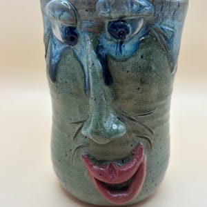May include: A ceramic mug featuring a sculpted face with a green speckled glaze, prominent nose, and deep blue accents around the eyes. The mug has a wide, open mouth with glossy red lips and carved lines around the nose and mouth area.