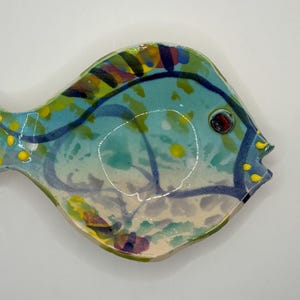 May include: A fish-shaped ceramic dish featuring a light blue base with hand-painted abstract patterns in dark blue, green, and yellow. The design includes a single dark red eye, yellow dots along the tail and mouth, and dark blue outlines.