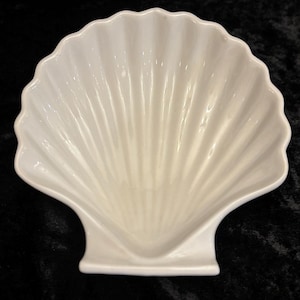 May include: A white ceramic seashell-shaped dish. The dish has a scalloped edge and a ribbed interior, resembling a real seashell. The dish is set against a dark background.