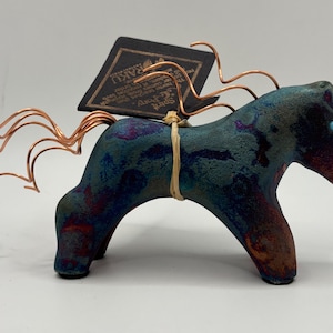 May include: A raku ceramic horse figurine with a textured teal and dark blue finish, featuring copper wire mane and tail accents. A tan raffia cord secures a black rectangular tag with the text Spirit Pony Raku to the horse's back.