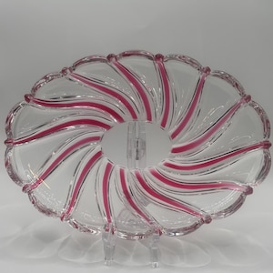 May include: Clear glass serving dish with a scalloped edge and a spiral design of pink stripes. The dish is oval-shaped and sits on a clear stand, showcasing its elegant design. A decorative piece for serving food.