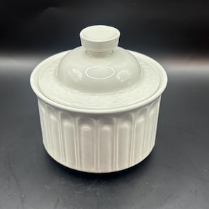 Puede incluir: Un azucarero de cerámica blanca con tapa. El cuenco tiene forma cilíndrica con ranuras verticales, y la tapa tiene un pequeño pomo. La tapa tiene un diseño decorativo. El azucarero está sobre una superficie negra.