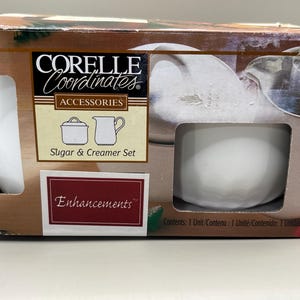 Pode incluir: Um conjunto de açucareiro e cremeira Corelle Coordinates na embalagem original. A caixa é castanha com uma ilustração de um açucareiro e um jarro de creme brancos. As palavras "Sugar & Creamer Set" estão impressas abaixo.