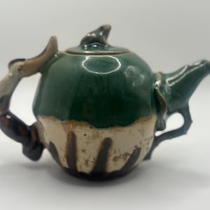 May include: A ceramic teapot featuring a glossy dark green upper section and a cream-colored lower base with vertical brown stripes. The lid is topped with a small sculpted frog figure, and the handle and spout have organic, textured shapes.