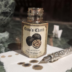 Puede incluir: Una pequeña botella de vidrio de tinta "Crow's Clutch" Black Walnut con una etiqueta con ilustraciones de nueces. Una pluma plateada y manchas de tinta están sobre una superficie blanca. Un mortero y una maja de mármol están en el fondo.