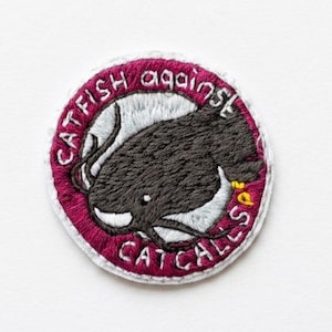 Könnte beinhalten: Gestickter Aufnäher mit einer dunkelgrauen Wels-Illustration in einem weißen Kreis, umrahmt von einem weinroten Rand. Die Worte "CATFISH against CAT CALLS" sind in Weiß gestickt. Der Aufnäher hat einen weißen Rand.