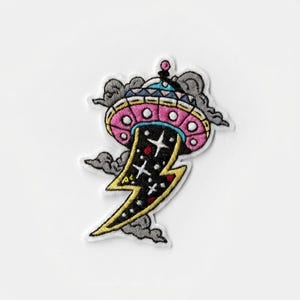Könnte beinhalten: Gestickter Aufnäher mit einem rosa und blauen UFO mit einem schwarzen und gelben Blitzdesign. Das UFO hat ein detailliertes Design mit grauen Wolken und Sternen. Der Aufnäher befindet sich auf weißem Hintergrund.