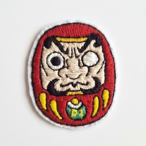 Könnte beinhalten: Gestickter Aufnäher mit einem Daruma-Puppen-Design. Der Aufnäher hat einen roten Hintergrund mit schwarzen, gelben und beigen Details. Die Daruma-Puppe hat ein offenes Auge und einen strengen Ausdruck. Der Aufnäher ist mit weißen Nähten umrandet.