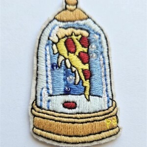 Puede incluir: Parche bordado que representa una porción de pizza dentro de un globo de nieve. La pizza tiene corteza amarilla, pepperoni rojo y está encerrada en una cúpula de cristal azul y transparente. La base y la parte superior del globo son doradas.