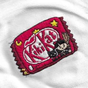 May include: Embroidered patch featuring a red Kit Kat candy bar wrapper design with white text and a cartoon character. The patch has a scalloped edge and yellow star and crescent moon accents, set against a white fabric background.