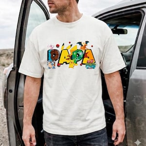 May include: Cream-colored t-shirt with the word "PAPA" in colorful letters, featuring cartoon characters. The shirt has short sleeves and a crew neck. The design includes various Pokemon characters.