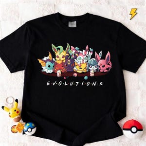 May include: Black t-shirt with a vibrant graphic of cartoon creatures and the word "EVOLUTIONS" in white. A Pikachu keyring, a Poke Ball, and other accessories are visible in the image.