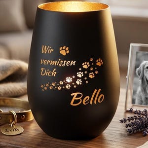 Può includere: Portacandele nero con interno dorato, con la scritta "Wir vermissen Dich Bello" e impronte di zampe. Sono visibili anche un collare dorato per cani e una foto incorniciata di un cane.