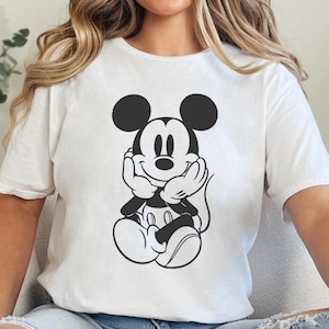 Peut inclure: T-shirt blanc avec un dessin noir et blanc de Mickey Mouse. Le personnage de dessin anim&eacute; est assis, les mains sous le menton. Le t-shirt est port&eacute; avec un jean bleu clair.