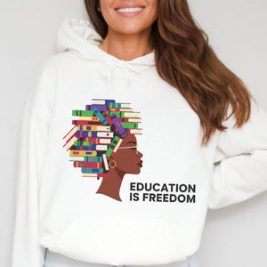 Puede incluir: Sudadera con capucha blanca con un gr&aacute;fico de un perfil humano con el pelo formado por una pila de libros coloridos. El texto "EDUCATION IS FREEDOM" est&aacute; impreso debajo. La sudadera tiene un bolsillo delantero y una capucha con cord&oacute;n.