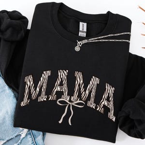 May include: Black sweatshirt with the word "MAMA" in a zebra print design. A silver necklace with a circular pendant rests on the sweatshirt. The sweatshirt is paired with blue jeans.