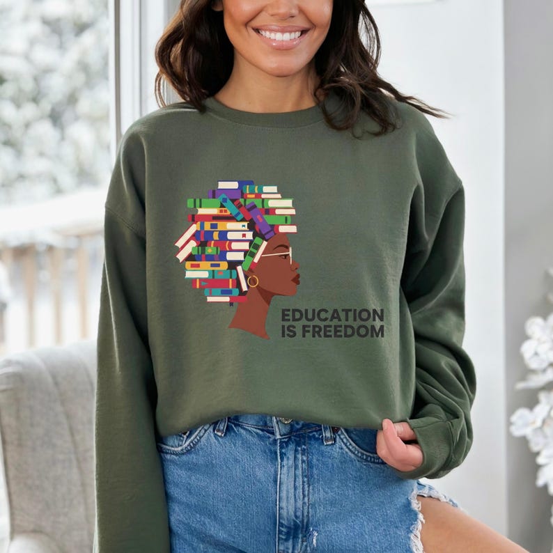 Puede incluir: Sudadera verde oliva con un gr&aacute;fico del perfil de una mujer con el pelo peinado con libros, un pendiente de aro dorado y gafas. El texto "EDUCATION IS FREEDOM" est&aacute; impreso en negro. La sudadera se combina con vaqueros azules.