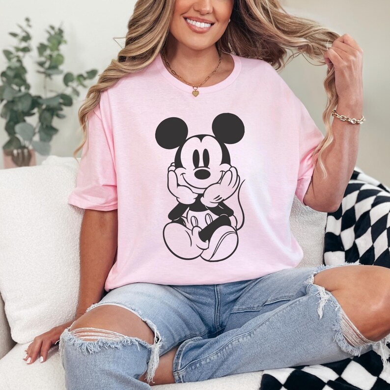 Peut inclure: T-shirt rose clair avec un dessin noir et blanc de Mickey Mouse. Le personnage de dessin anim&eacute; est assis, les mains sous le menton. Le t-shirt a des manches courtes et un col rond.
