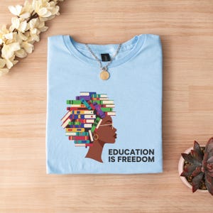 Puede incluir: Camiseta azul claro con un gr&aacute;fico del perfil de una mujer, con el pelo peinado como una pila de libros coloridos. El texto "EDUCATION IS FREEDOM" est&aacute; impreso debajo de la imagen. Un collar con colgante dorado est&aacute; sobre la camiseta.