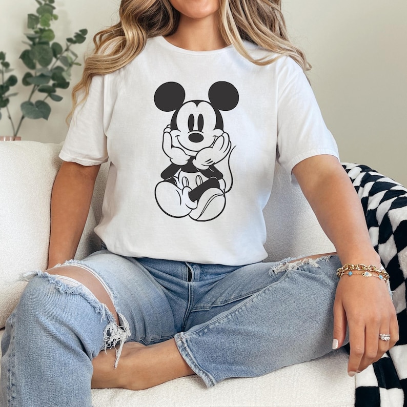 Peut inclure: T-shirt blanc avec un dessin noir et blanc de Mickey Mouse. Le personnage de dessin anim&eacute; est assis, les mains sous le menton. Le t-shirt est port&eacute; avec un jean bleu clair.