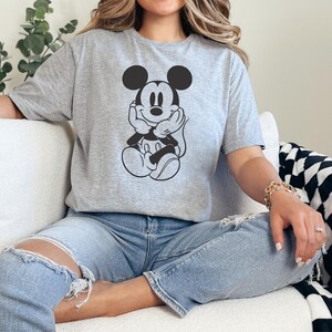 Peut inclure: Un t-shirt gris avec un dessin noir et blanc de Mickey Mouse. Le personnage de dessin anim&eacute; est assis, les mains sous le menton. Le t-shirt est port&eacute; avec un jean bleu.